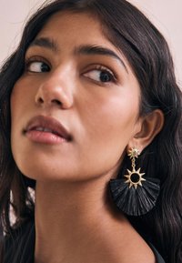 Black fan-shaped earrings with a gold sun accent, featuring a circular black center, textured material, and detailed hardware.