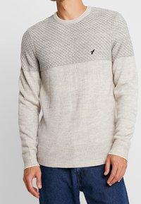 Maglione grigio chiaro a maglia con un motivo a diamante testurizzato sulle spalle e polsini e orlo a costine. Presenta un sottile accento con logo.