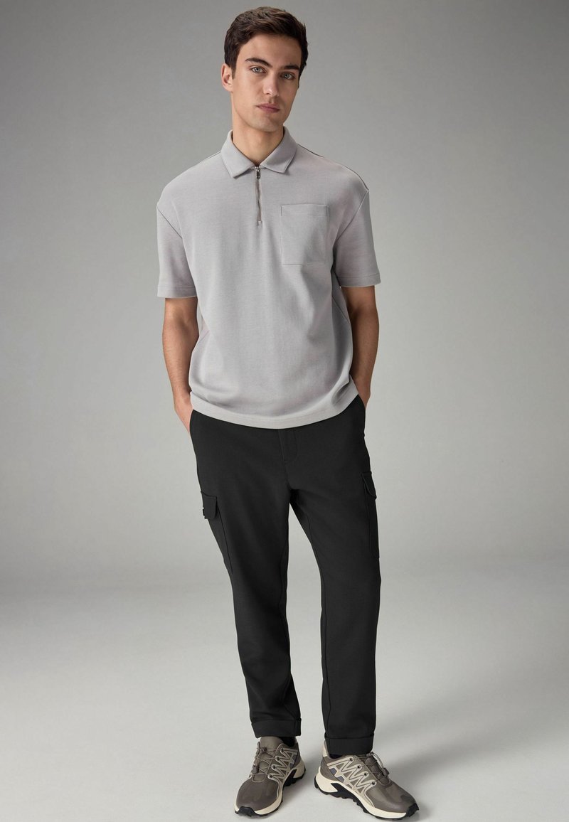 Polo de cremallera gris claro con mangas cortas y un bolsillo, combinado con pantalones cargo negros con bolsillos laterales y zapatillas grises.