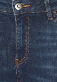 Pantalones vaqueros de mezclilla azul oscuro con costuras naranjas. Incluyen un cierre de botón y dos bolsillos delanteros con una textura ligeramente descolorida.