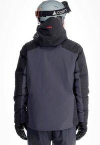 Persona che indossa una giacca invernale nera e grigio scuro con cappuccio, guanti neri e un casco Cairn grigio vista di spalle su sfondo bianco.