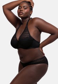 Juodas nėrinių liemenėlė su gilia iškirpte ir reguliuojamomis petnešėlėmis, derinama su atitinkamais nėrinių apatiniais, turinčiais banguotus kraštus.