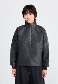 Svart zip-up jacka med hög krage, med en glansig finish, vinkelartad design och sidofickor. Nike-logotyp på bröstet.
