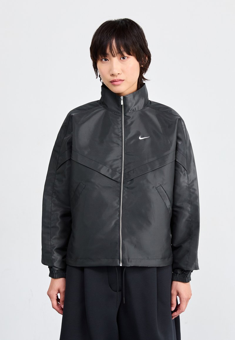 Svart zip-up jacka med hög krage, med en glansig finish, vinkelartad design och sidofickor. Nike-logotyp på bröstet.