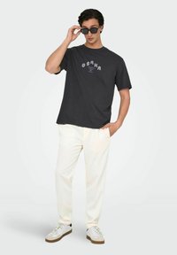Sort bomulds T-shirt med "OSAKA" grafik i lilla, parret med cremefarvede bukser og hvide sneakers, model der justerer mørke solbriller.