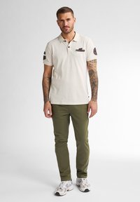 Béžová polo tričko s límečkem a dvěma knoflíky, s malou nášivkou na hrudi a olivově zelenými slim-fit kalhotami. Bílé tenisky.