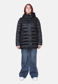 Zwarte pufferjack met capuchon, met gestikte delen en een rits aan de voorkant. Draagt over loszittende denim broek en witte sneakers.