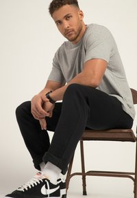 Grå bomullströja, svarta sweatpants och svarta sneakers med vita detaljer. Sitter på en metallstol, avslappnad hållning med händerna sammanflätade.