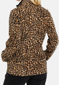 Kobieta w skórzanym płaszczu w cętki, z długimi rękawami, stojąca z rękami w kieszeniach, tyłem do aparatu.