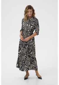 Lång, knäppt klänning i zebramönster, med neutrala nyanser av beige och svart, och en bältesmidja samt volangkjol. Svarta skor.