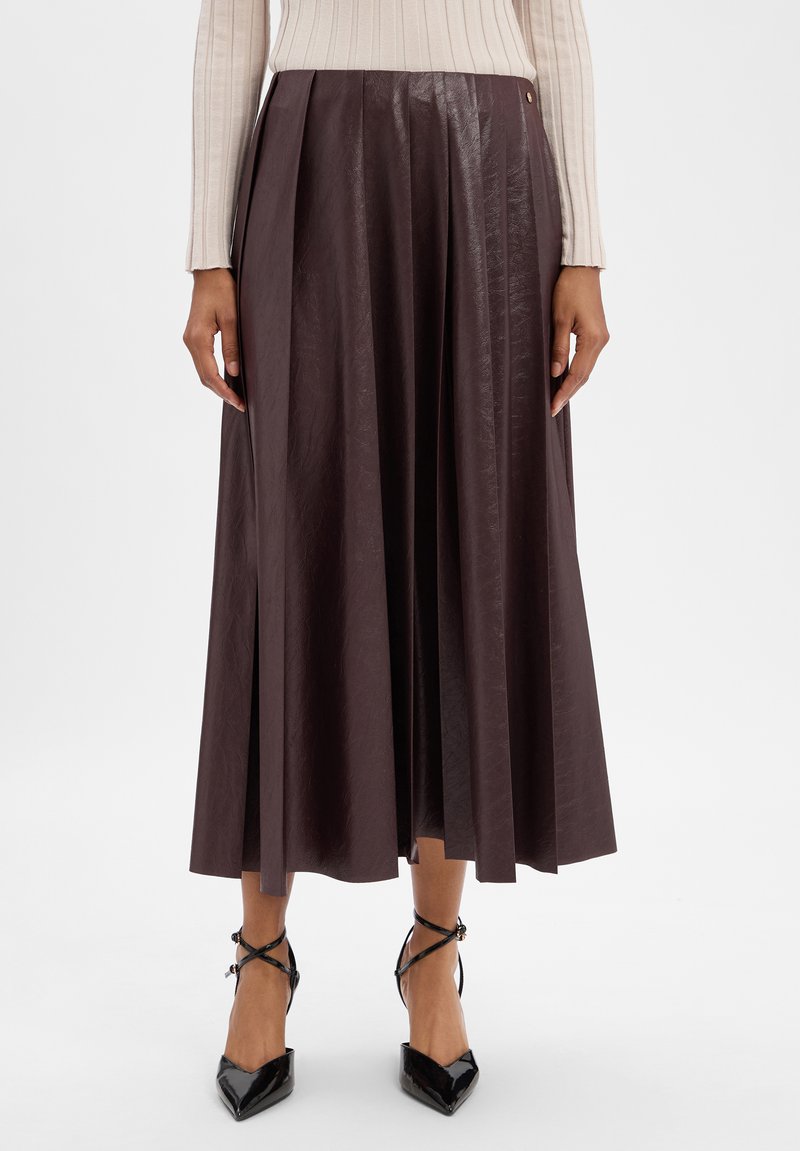 Femme portant une jupe midi plissée marron foncé, un haut beige à manches longues côtelé et des escarpins noirs à bout pointu avec des lanières à la cheville.