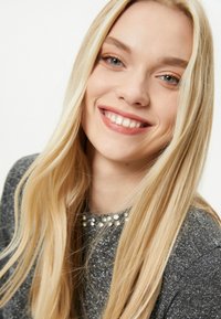 Szary sweter z błyszczącymi nitkami i kołnierzykiem zdobionym perłami, w połączeniu z długimi, prostymi blond włosami i promiennym uśmiechem.
