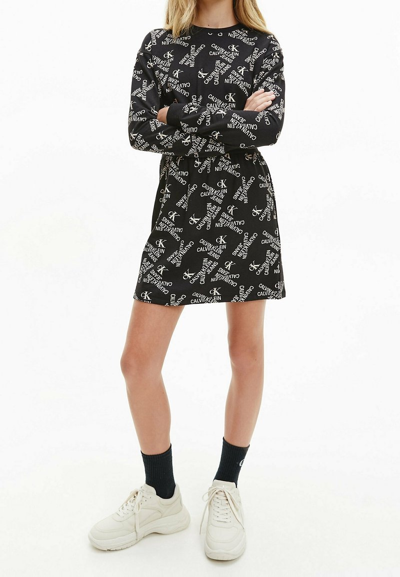 Femme portant une robe noire à manches longues avec des logos blancs Calvin Klein Jeans, des chaussettes noires et des baskets blanches, debout les bras croisés.