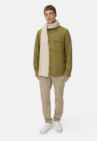 Gequilte olijfgroene jas met voorkant zak, gecombineerd met een crèmekleurige geribbelde sjaal, beige broek en witte sneakers, staand tegen een witte achtergrond.