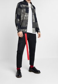 Chemise à motifs noirs et gris à boutons, t-shirt blanc en dessous, pantalon cargo noir avec sangle accent rouge, chaussures noires et chaussettes rouges.