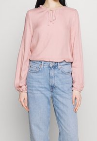 Blusa rosa a maniche lunghe con scollatura volant e dettagli legati sul davanti, realizzata in un tessuto liscio, abbinata a jeans blu a vita alta.