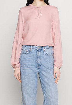 Blusa rosa a maniche lunghe con scollatura volant e dettagli legati sul davanti, realizzata in un tessuto liscio, abbinata a jeans blu a vita alta.