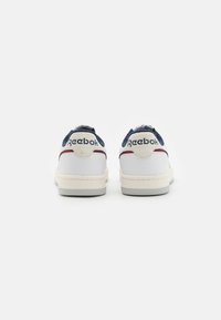 Vit sneaker med marinblå detaljer, med en slät läderövre, rund tå, kontrasterande röd rand och Reebok-logotyp på hälen.