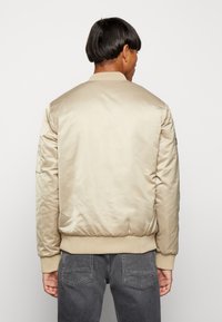 Giacca bomber beige con una finitura liscia e lucida, polsini e orlo a costine, e una toppa sul braccio sinistro. Indossata con pantaloni grigi.