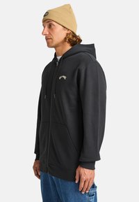 Schwarze Zip-Hoodie mit einer Fronttasche, gerippten Bündchen und einem kleinen Logo auf der Brust; kombiniert mit einer beigen Strickmütze.