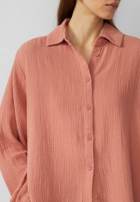 Donna che indossa una camicia button-up testurizzata di colore rosa salmone con colletto, vista dalla spalla al mento, su uno sfondo chiaro e semplice.