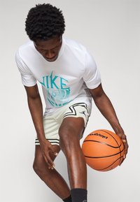 Hvid Nike t-shirt med turkist logo, båret sammen med lette, mønstrede shorts. Spilleren holder en orange basketball og viser dynamisk bevægelse.