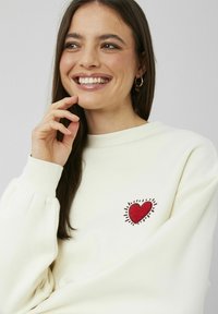 Sweatshirt couleur crème avec un cœur rouge brodé et des accents de rayons de soleil sur la poitrine ; tissu tricoté avec des poignets côtelés.
