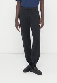 Pantalones deportivos negros hechos de tela suave, con cintura elástica con cordón, puños acanalados y un ajuste holgado, combinados con zapatos negros sin cordones.