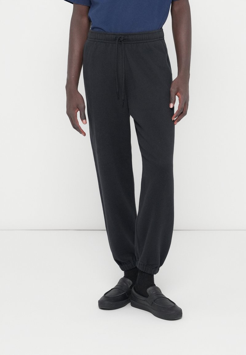 Pantalones deportivos negros hechos de tela suave, con cintura elástica con cordón, puños acanalados y un ajuste holgado, combinados con zapatos negros sin cordones.