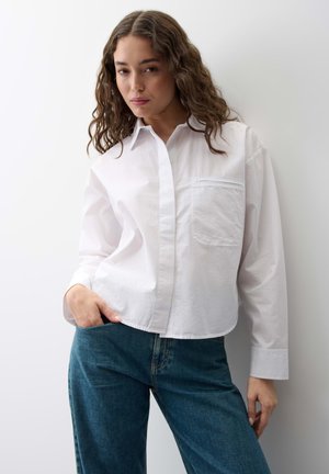 Giovane donna con capelli ondulati che indossa una camicia bianca larga con bottoni e jeans blu, in piedi davanti a uno sfondo chiaro e semplice.