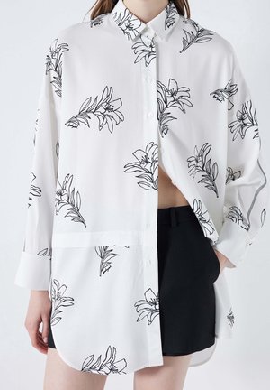 Weißes langärmliges Hemd mit schwarzen floralen Umrissen, ungeknöpft über schwarzen Shorts getragen an einer Person mit einer Hand in der Tasche.