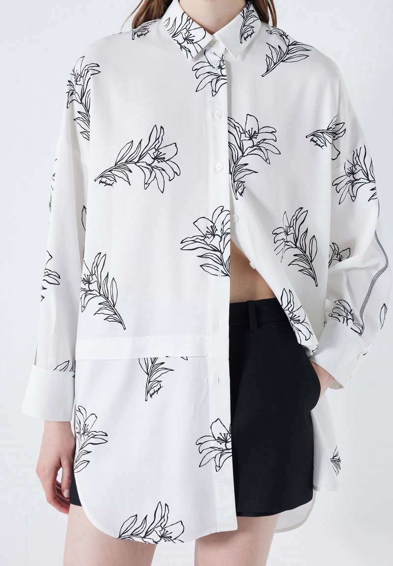 Chemise blanche à manches longues boutonnée avec des contours floraux noirs, portée non rentrée sur un short noir sur une personne avec une main dans la poche.