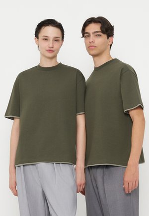 Deux jeunes adultes portant des chemises à manches courtes vert olive et des pantalons gris clair se tiennent côte à côte devant un fond blanc uni.