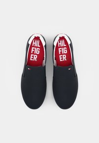 Paire de chaussures noires sans lacets avec semelles blanches, ornées de semelles intérieures rouges imprimées avec "HILFIGER" et d'un petit drapeau Tommy Hilfiger sur chaque chaussure.