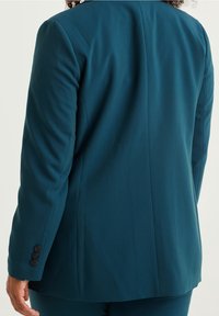 Blågrön kavaj med figursydd design, tillverkad av slätt tyg, med en bakre söm och treknappsmuddar. Rent, skräddarsytt utseende.