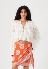 Hvit bluse med rynker og lange ermer, med knyting, kombinert med en oransje omslagskjørt med hvite paisleymønstre.