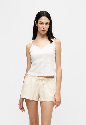 Femme aux longs cheveux foncés portant un haut blanc sans manches côtelé et un short beige clair avec un petit motif floral et une taille à cordon.