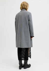Manteau en mélange de laine gris avec un col tailleur, silhouette droite et manches longues, associé à une jupe en dentelle noire et des bottes noires jusqu'aux genoux.