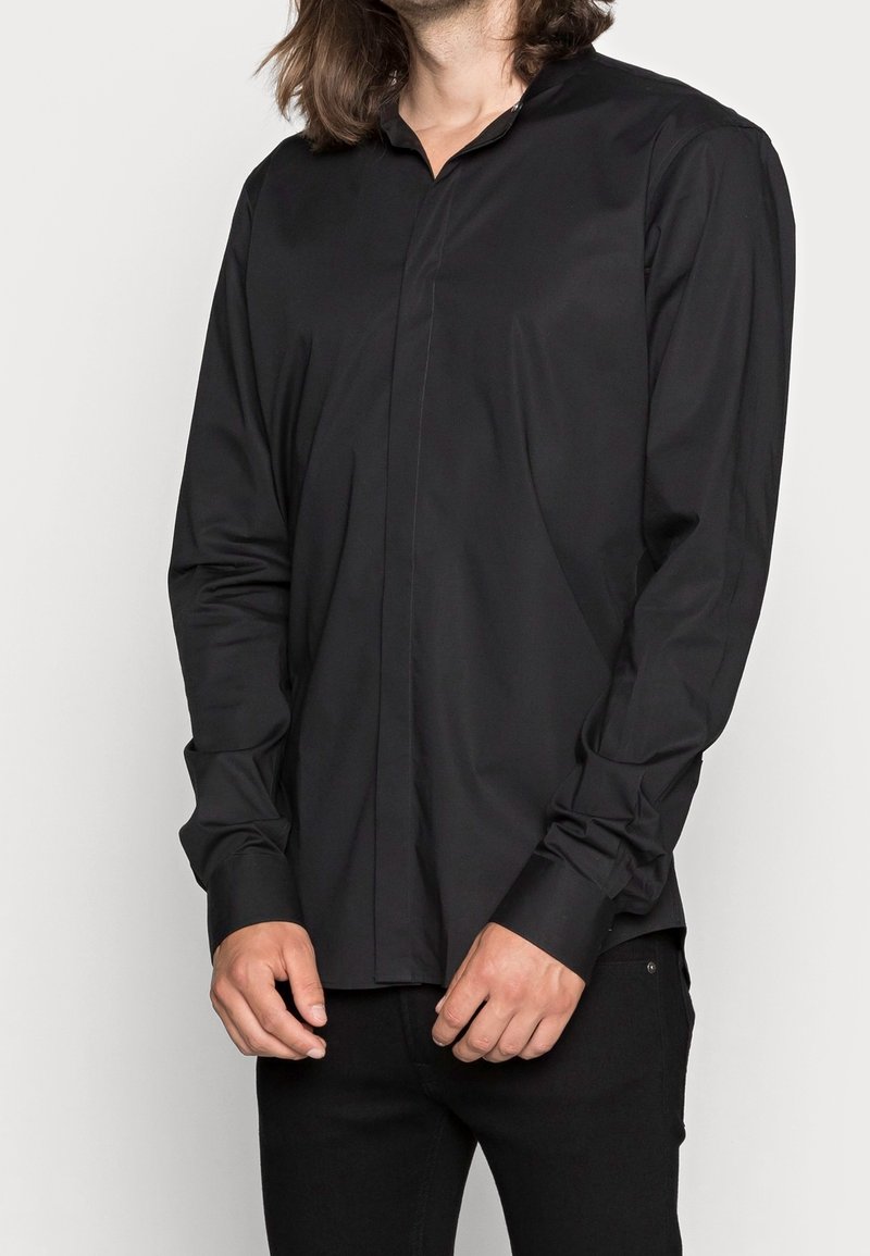 Personne portant une chemise noire à manches longues boutonnée et un pantalon noir, debout les mains devant sur un fond uni.