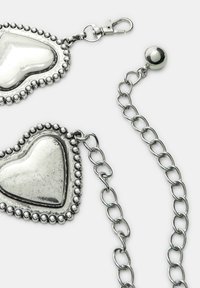 Srebrne kolczyki w kształcie serca z koronkowym wykończeniem, mocowane za pomocą zapięcia i połączonego łańcuszka, z polerowanym metalicznym wykończeniem.