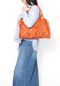 Sac à main en cuir orange avec de multiples poches, quincaillerie métallique et un design structuré. Porté à l'épaule par une personne en tenue décontractée.