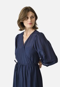 Marineblaues Wickelkleid mit Puffärmeln, aus strukturiertem Stoff und mit einem Taillengurt für eine anpassbare Passform. Dezentes gewebtes Muster sichtbar.