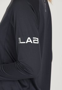 Pitkähihainen, tummansininen urheilusormi, jossa on teksturoitu kangas. Ominaisuuksina valkoinen "LAB"-logo hihassa ja ilmanvaihtoyksityiskohdat sivuilla.
