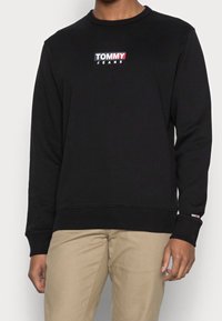 Sudadera negra de manga larga, con puños y dobladillo acanalados, que presenta un colorido logo de "TOMMY JEANS" en el pecho. Textura suave, ajuste casual.