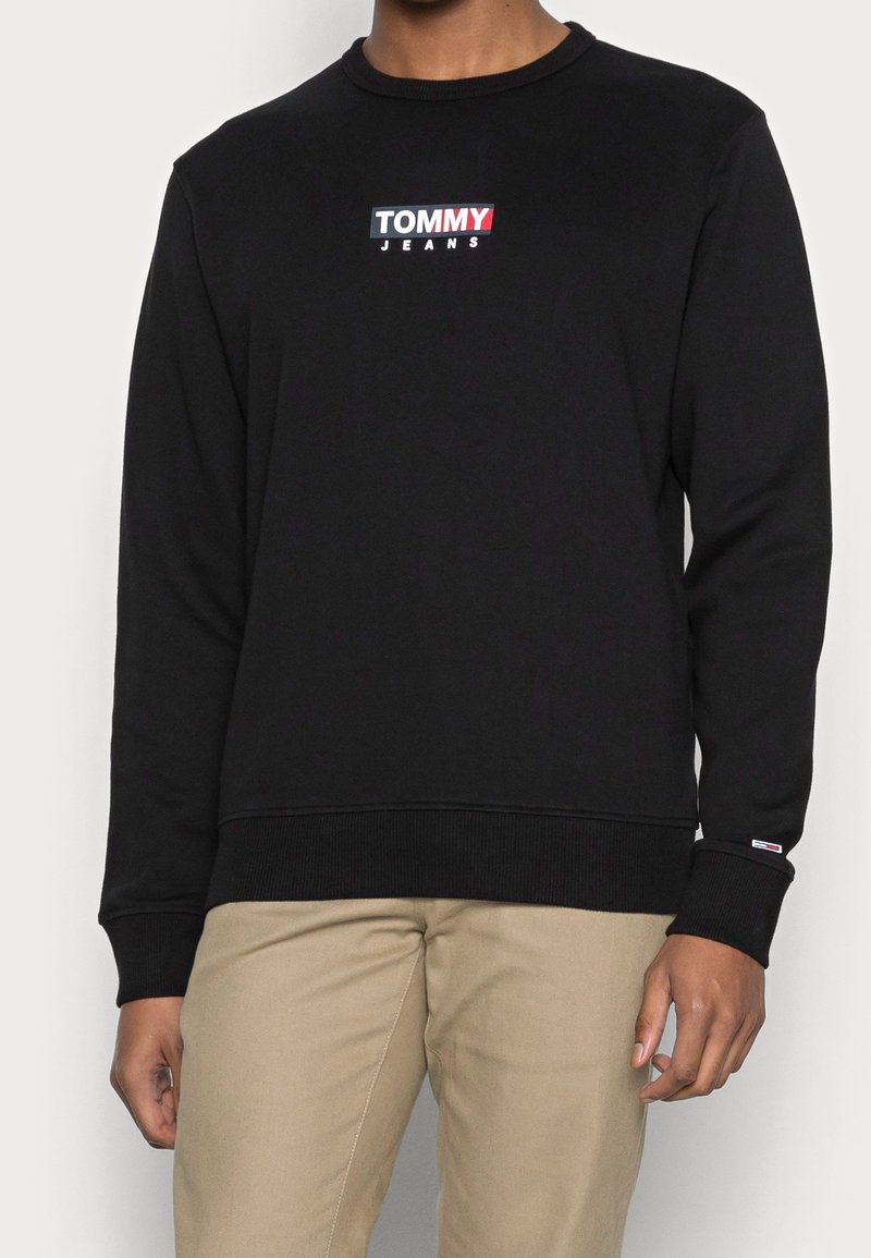 Sudadera negra de manga larga, con puños y dobladillo acanalados, que presenta un colorido logo de "TOMMY JEANS" en el pecho. Textura suave, ajuste casual.