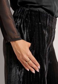 Zwarte fluwelen broek met verticale ribbelstructuur en een elastische tailleband, gecombineerd met een doorschijnende top met lange mouwen. Accentring op een hand.