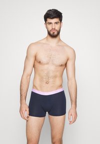 Boxer-strings en coton noir avec une ceinture élastique rose clair portant le texte "TOMMY HILFIGER". Texture lisse, design ajusté, longueur au-dessus du genou.