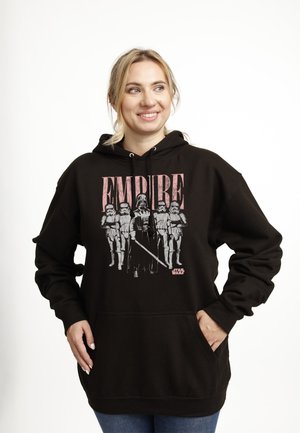 Uśmiechnięta kobieta w czarnej bluzie z kapturem z grafiką Star Wars Empire z Darthem Vaderem i szturmowcami, ręce w kieszeniach, stojąca na białym tle.