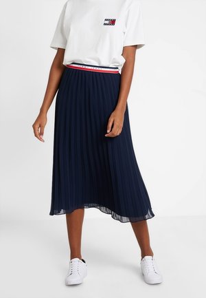 Femme portant un t-shirt blanc avec logo, une jupe midi plissée bleu marine avec une ceinture de marque, et des baskets blanches, debout devant un fond blanc uni.