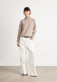 Donna che indossa un maglione beige a collo alto, pantaloni bianchi a gamba larga con una cintura nera e scarpe beige, in piedi davanti a uno sfondo bianco uniforme.